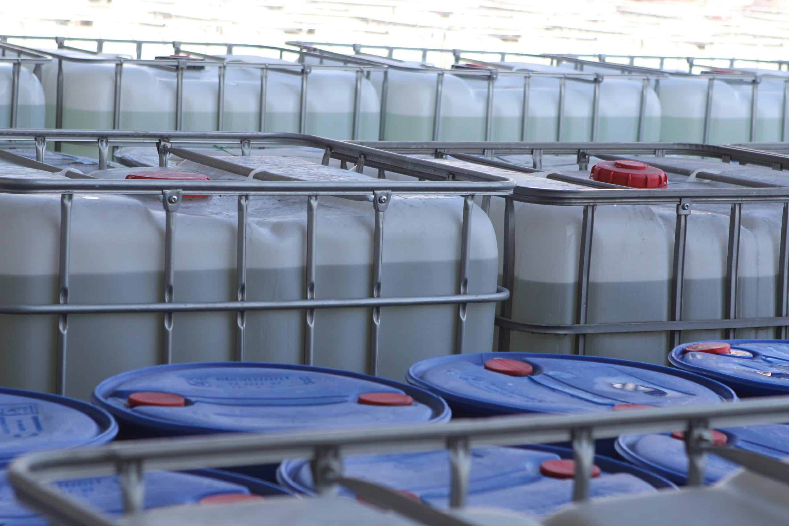 Rows of large white industrial liquid storage containers (IBC totes) inside a facility, each partially filled with light-colored liquid and secured in metal frames, with blue barrels visible in the foreground and red caps on the containers.